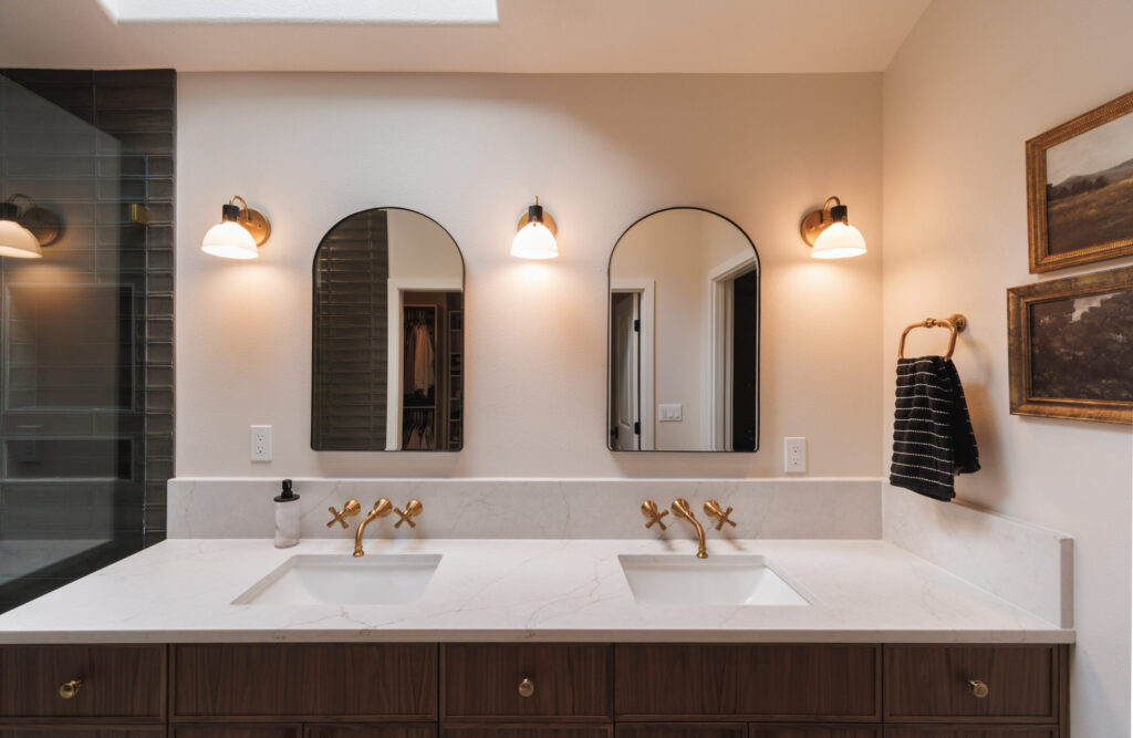 A primary bathroom remodel featuring a vanity with double sinks and mirrors, 3 vanity lights in brushed gold, and both faucets in the same brushed gold. The countertops are quartz. 