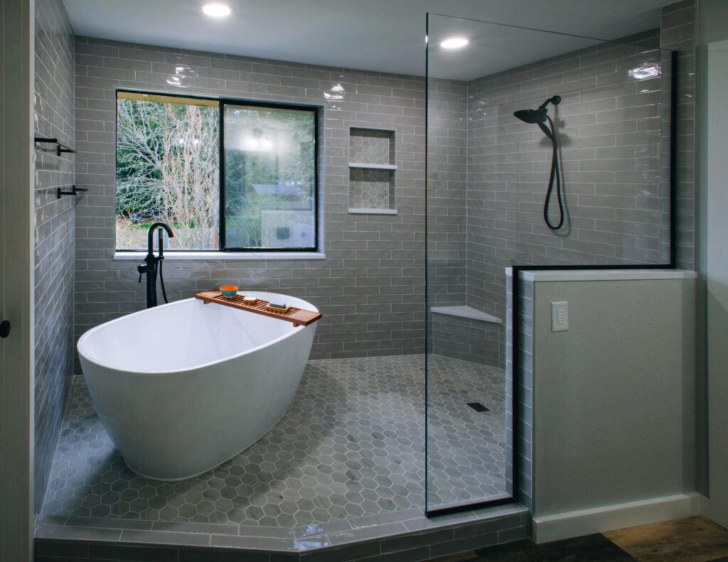 A bathroom remodel with large wet room equipped with a freestanding tub, grey subway tile on the walls, stone hexagon tile on the floor, a freestanding tub and black shower fixtures. 